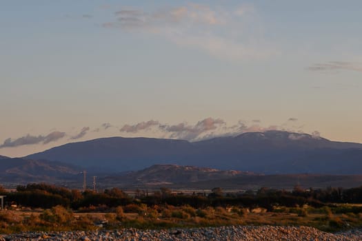 Golden hues from the sunset bathe the mountains in warm light, as soft clouds drift above the serene landscape. Nature's beauty unfolds in a tranquil evening setting that captivates the viewer.