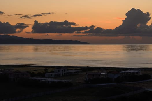 At dusk, the tranquil coastline is illuminated by a stunning sunset. Soft colors blend as gentle waves lap against the shore and quiet houses sit, silhouetted against the sky.