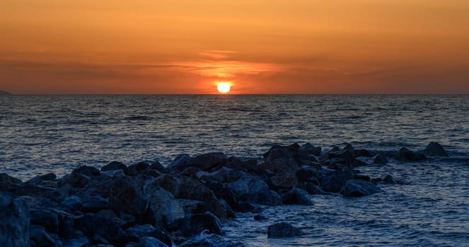 Warm hues of sunset blend seamlessly over the calm sea, creating a picturesque moment. Rocks in the foreground accentuate the serene setting, inviting tranquility and reflection.