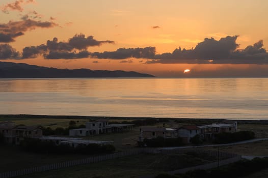 Soft hues of orange and pink paint the sky as the sun descends into the horizon. Peaceful waters reflect the colors, while quiet homes stand sentinel along the coast, enhancing the serene atmosphere.