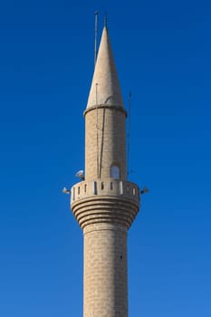 A striking minaret rises gracefully, showcasing intricate stonework and a tall spire. Nestled in a lively town, it stands as a testament to rich cultural heritage under a bright blue sky.