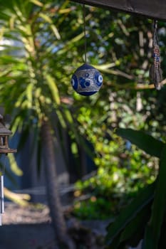 A vibrant ceramic ornament dangles from a branch, catching sunlight amid a backdrop of green leaves. The garden exudes tranquility, inviting peaceful moments outdoors.