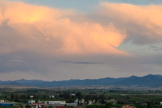 As dusk approaches, enchanting clouds fill the sky, painting it in pastel shades over the distant mountains. Below, lush fields extend towards a peaceful horizon, fostering a sense of calm.