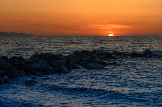 As the sun descends towards the ocean, vibrant colors fill the sky. Waves gently lap against the rocky coastline, offering a serene view of nature's beauty at twilight.