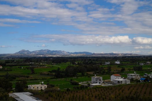 Lush green fields stretch across the horizon, dotted with charming residences. Majestic mountains rise in the distance beneath a bright blue sky with fluffy clouds.