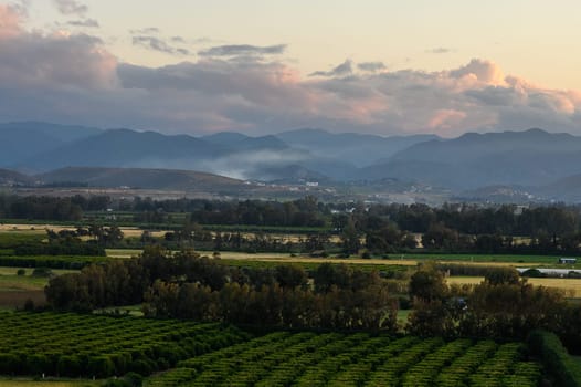 A beautiful expanse of lush green fields stretches across the countryside under a stunning sunset sky. Gentle hills rise in the background, creating a serene atmosphere as day transitions to night.