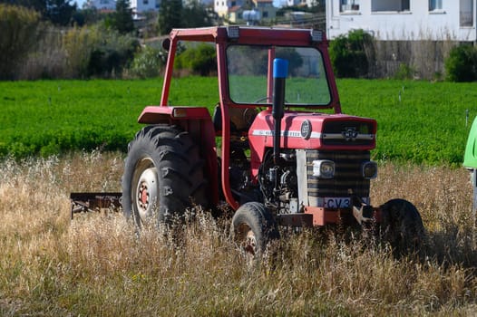 Gaziveren Cyprus -10.04.2025 An abandoned red tractor sits in a lush green field surrounded by golden grass. The bright afternoon sun casts shadows, revealing the beauty of rural life and nature's harmony.