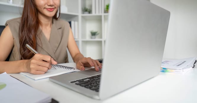 A woman confidently jots down notes as she engages in her work on a laptop, showcasing an organized and productive work atmosphere.