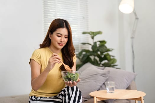 Emphasizing nutrition, a young woman savors a fresh green salad, focusing on healthy eating habits that contribute to her well-being in her cozy living space.