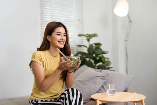 A cheerful woman sits on the couch, savoring a nutritious salad in the bright morning light, embracing a healthy start to her day.