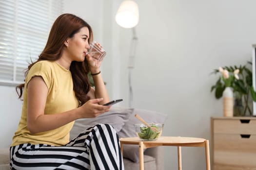 A woman balances hydration and nutrition by sipping water next to her healthy salad, fully embracing her refreshing morning routine.