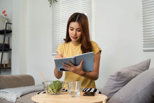 Focused on her health journey, a young woman writes in her notebook while savoring a nutritious salad in a cozy home setting, reflecting her commitment to wellness.