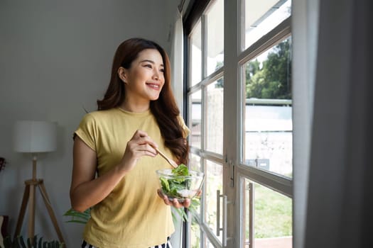 A young woman stands by the window, smiling while enjoying a vibrant bowl of salad, embodying the essence of mindful eating and appreciation for nutritious food.