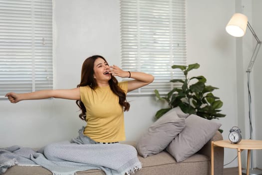 A young woman wakes up, stretching and yawning in a cozy living room, embracing the morning with positivity.