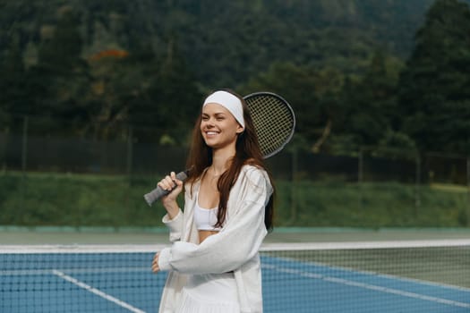 A joyful young woman enjoys playing tennis outdoors in a stylish white sports outfit, radiating confidence with a bright smile on a sunny day amidst lush greenery