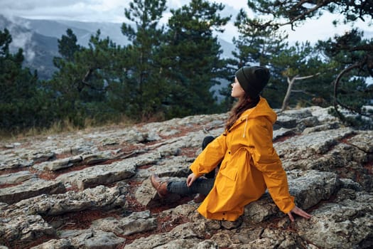A woman in a stylish yellow raincoat sitting peacefully in front of a majestic mountain, embracing the beauty of nature