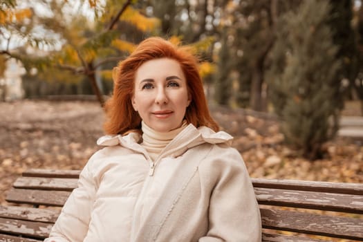 Red-haired woman in a cream jacket sits on a park bench, autumn leaves scattered around. She looks directly at the camera.