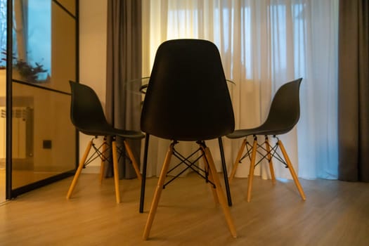 Three black Eames-style chairs surround a small dining table in a brightly lit room with sheer curtains and a mirrored partition.