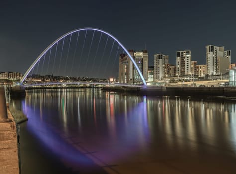 Newcastle, UK - Apr 13, 2025 - Nightscape scenery of The Iconic gateshead millennium bridge spanning the River Tyne and Baltic flour mills with Dark sky in the background. The pedestrian bridge with reflection in the river, use it as your Wallpaper, Poster and Space for text, Selective focus.