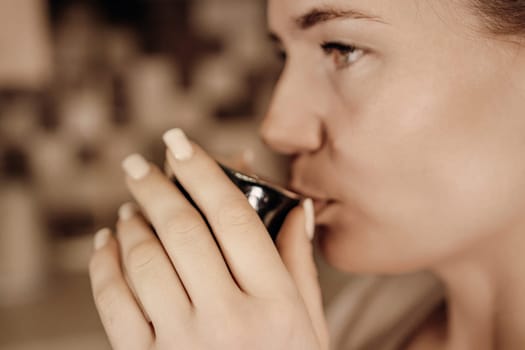 Close-up profile of a woman with light skin and manicured nails delicately sipping from a small, dark-colored cup.