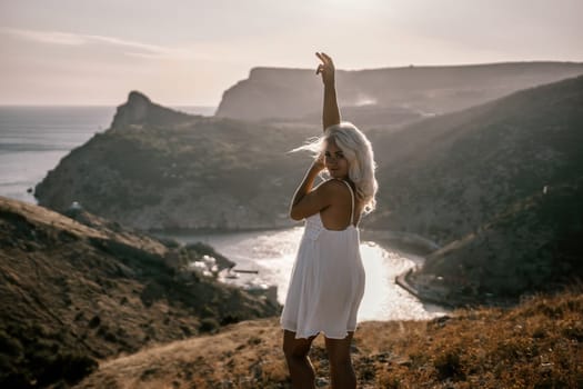A woman stands on a hill overlooking a body of water. She is wearing a white dress and she is enjoying the view