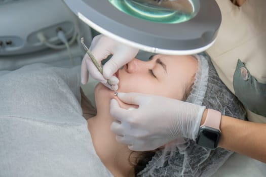 Close-up of a woman receiving a facial treatment. A technician uses a small tool to cleanse her skin under a magnifying lamp. Gloves and a hygienic head covering are visible.