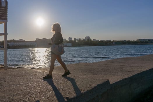 A woman walks along a concrete pier towards the setting sun. The sun is shining brightly and the water is calm. The cityscape is visible in the background.