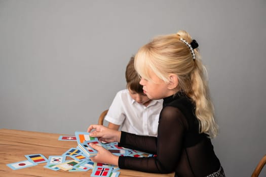 A young girl with blonde hair sits at a table, looking at a stack of cards, while a boy sits beside her, out of focus.