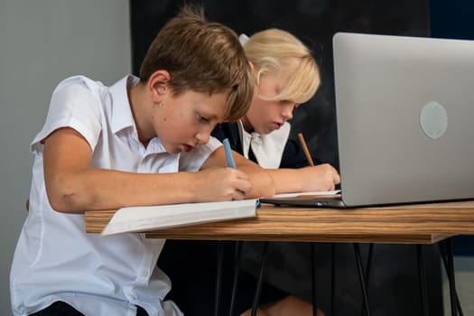 Two children in school uniforms sit at a table and write on paper while a laptop sits open in front of them.