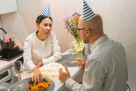 Here's a caption describing the image: A man and woman, wearing party hats, sit at a table. A birthday cake with lit candles is between them. They share a meal and drinks in a small, intimate setting.