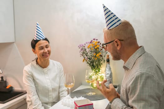 Here's a caption under 150 characters: A bald man and a woman, both wearing party hats, share a laugh and a drink at an intimate birthday celebration. A gift is present.