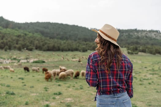 A woman wearing a straw hat stands in a field with a herd of sheep. The scene is peaceful and serene, with the woman looking out over the animals