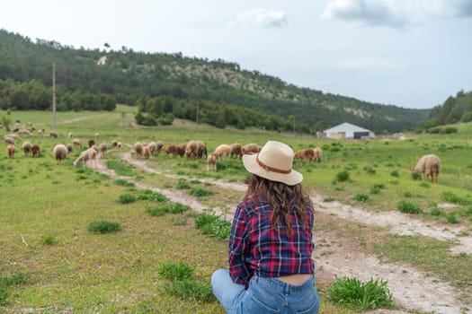 A woman in a plaid shirt and hat is sitting in a field with a herd of sheep