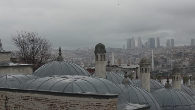 Overcast Istanbul skyline view from a rooftop. Grey domes, chimneys, and a sprawling cityscape beneath a cloudy sky. Modern skyscrapers visible in the distance.