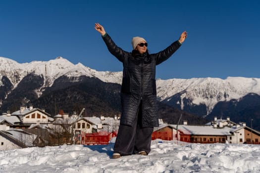Woman in long coat joyfully raises arms, overlooking snow-covered village nestled in majestic, snow-capped mountains under a vibrant blue sky.