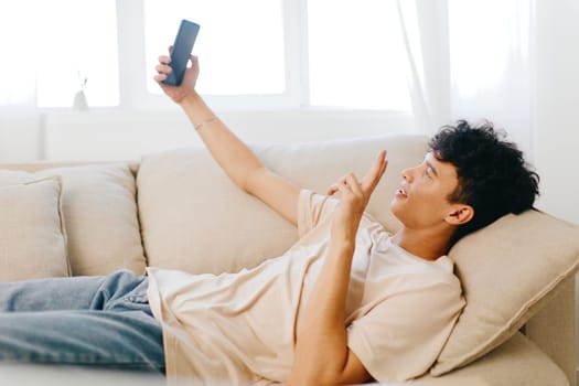 A young man is comfortably lying on a beige couch while holding a smartphone, engaging in a video call The warm, bright atmosphere creates an inviting home environment