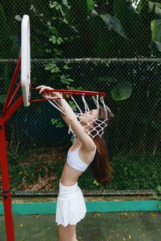 Energetic young woman joyfully celebrates her basketball success, hanging from the hoop She wears a white athletic outfit, surrounded by lush greenery on an outdoor court