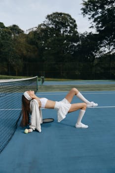 A young female tennis player wearing a stylish sports outfit enjoys a moment of relaxation on the court, exuding happiness and confidence in a sunny outdoor environment, ideal for summer sports