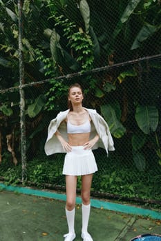 A confident young woman in a white sporty outfit stands on a tennis court, exuding a vibrant and energetic vibe amid lush green foliage The image reflects fitness, style, and a connection to nature