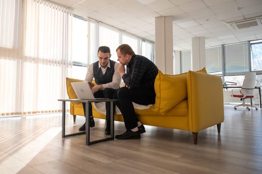 Two Caucasian men discussing work on a laptop while sitting on a sofa. Modern office