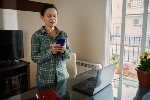 A woman stands in a home office, using a smartphone beside her laptop. The setting features a glass table, a notebook, and potted flowers, emphasizing a productive and casual atmosphere.