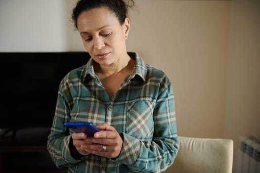 A woman is seen in a home environment using her smartphone, appearing thoughtful. She wears casual and comfortable attire, positioned in a domestic setting reflecting modern lifestyle habits.
