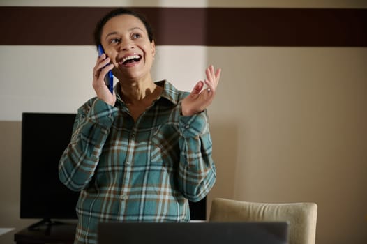 A joyous woman wearing plaid speaks on her smartphone in a home office environment. The scene conveys connection, warmth, and productivity, symbolizing a balanced modern lifestyle.
