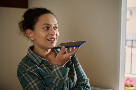 A woman uses a smartphone to record a voice message while in a cozy home environment. Her casual, relaxed demeanor conveys a personalized interaction with modern technology and communication tools.