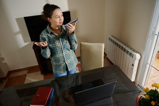 A young woman, dressed casually, speaks into her smartphone while standing at her home office desk. Surrounding items include a laptop, a notebook, and a cozy home interior, suggesting remote work.
