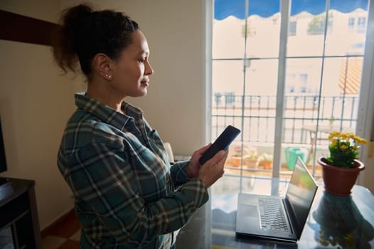 A focused woman in a home office setting uses her smartphone and laptop for remote work. The indoor ambiance, brightened by natural light, reflects dedication and modern telecommuting practices.
