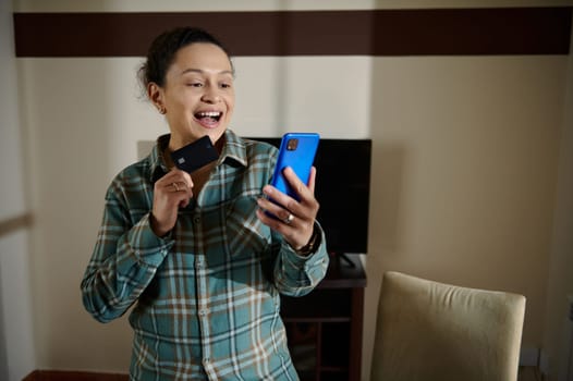 A cheerful woman using her smartphone and holding a credit card while shopping online. The interaction showcases modern digital payment methods and the convenience of e-commerce from home.