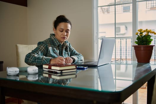 A concentrated woman working at her desk, taking notes on a journal next to her laptop. The surroundings exude a professional and organized vibe, featuring a serene indoor setting.