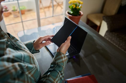 A person interacts with a smartphone while working on a laptop in a cozy home setting. The warm natural lighting and indoor plants contribute to a peaceful and focused workspace ambiance.