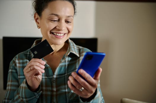 Woman smiling while using a smartphone and holding a credit card, representing online shopping and digital transactions. Remote connectivity and online payment convenience are evident.